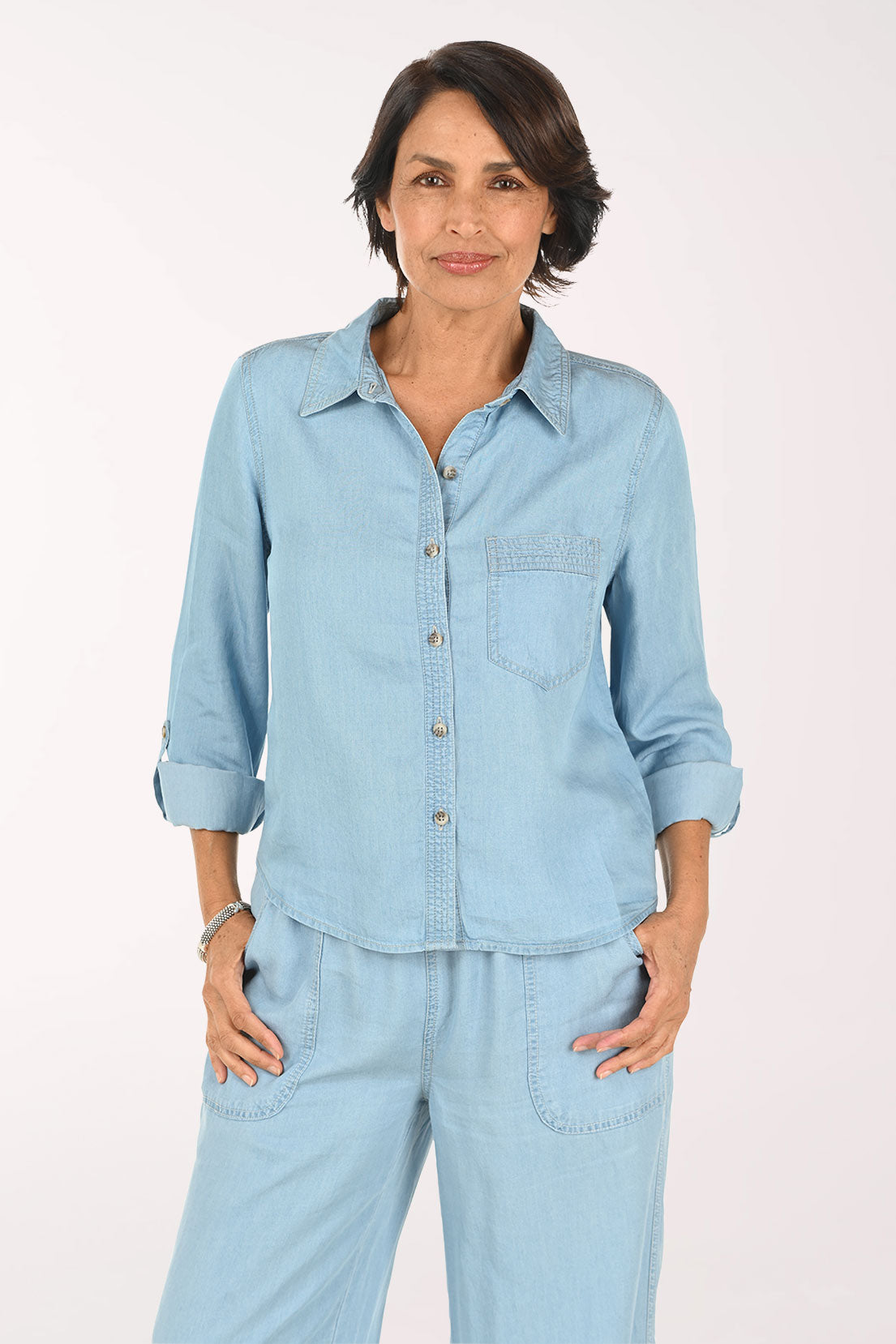 A smiling woman wearing a light blue denim button-up shirt and matching light blue denim pants, photographed against a plain light background.