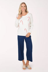 A woman wearing dark medium wash wide leg cropped pull-on jeans with a white sweater with red and white candy cane motifs and nude slide sandals.