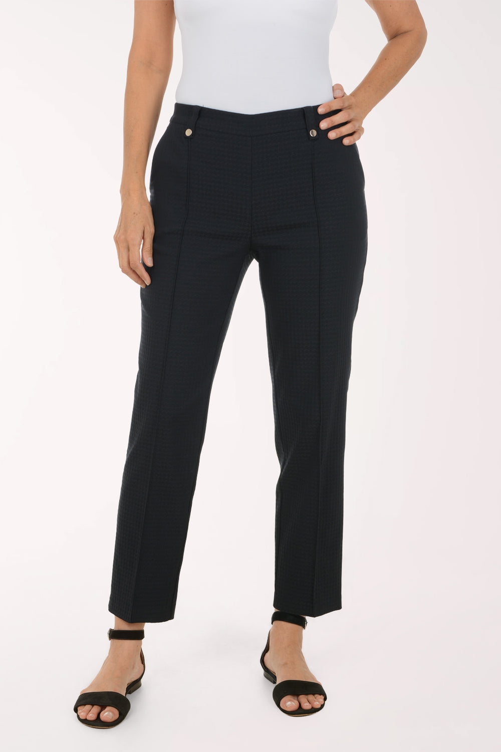 A model wearing black textured straight-leg trousers with gold-tone button details, paired with a white top and black sandals, shown from the waist down on a white studio background.