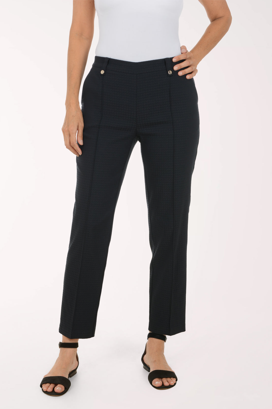 A model wearing black textured straight-leg trousers with gold-tone button details, paired with a white top and black sandals, shown from the waist down on a white studio background.