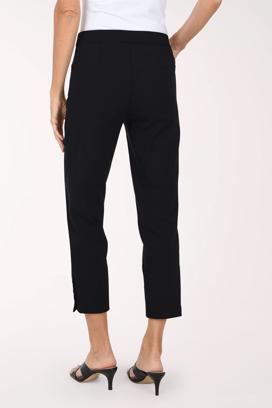 A person modeling black cropped pants with three decorative gold-tone buttons at the hem, paired with black mule sandals against a plain light background.