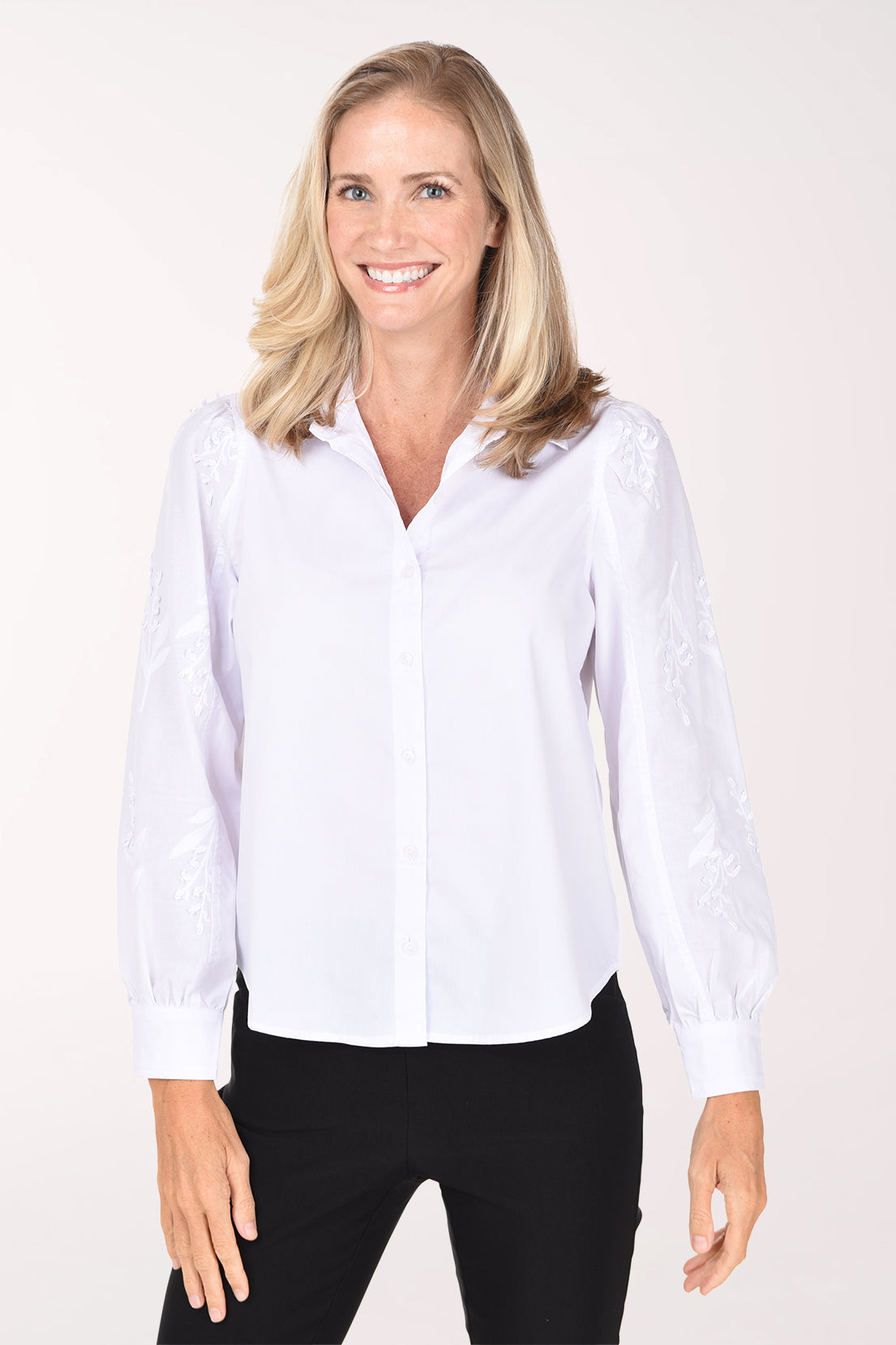 A woman wearing a white long-sleeve blouse with delicate floral embroidery on the sleeves and black pants, photographed against a plain white studio background.