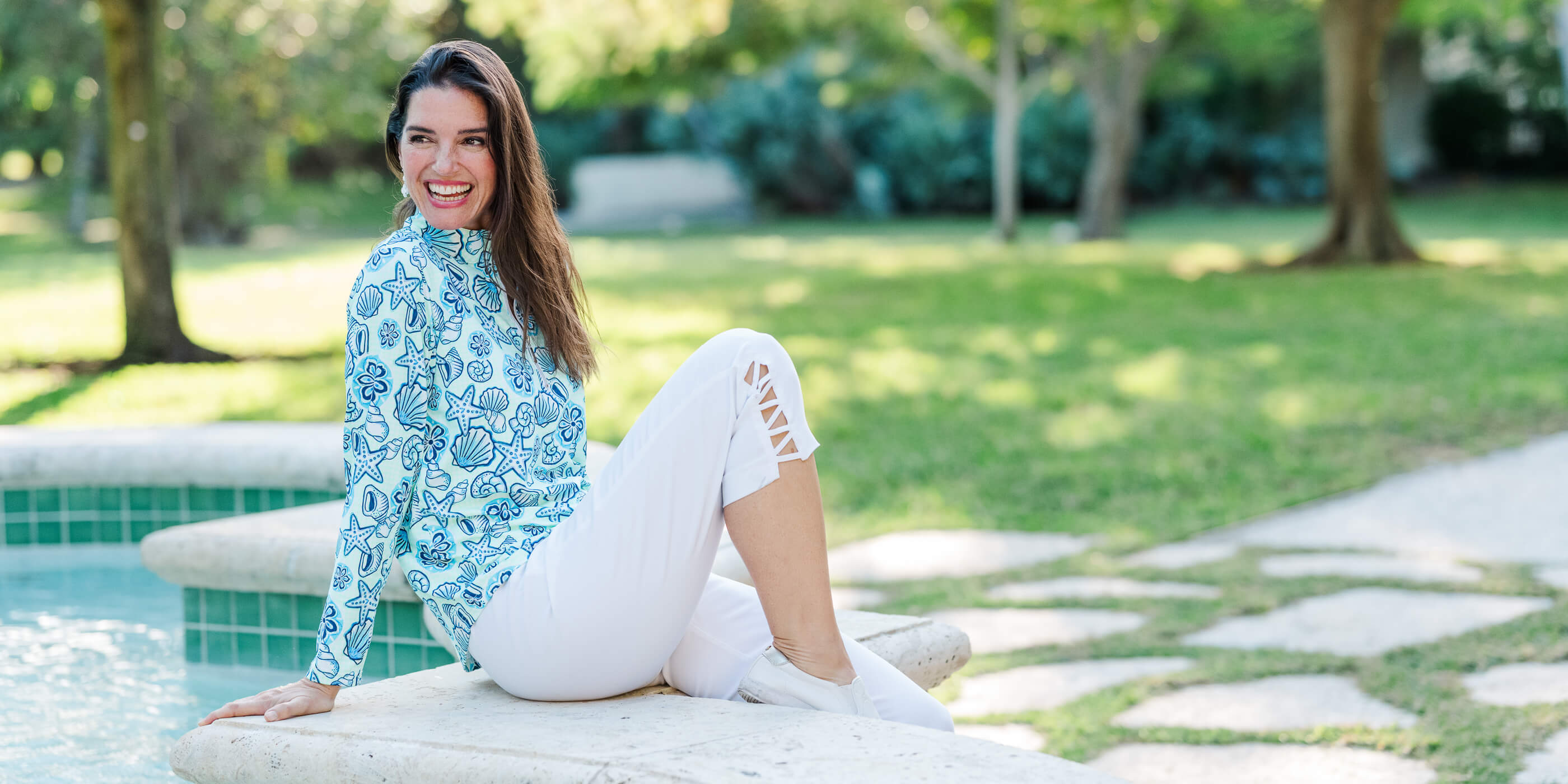 Woman sitting by a pool wearing a blue patterned top and white pants.