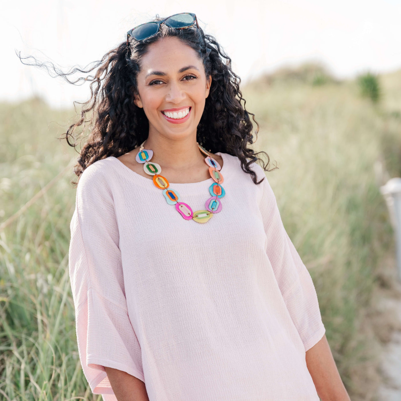 A smiling woman with curly dark hair wearing a light pink short-sleeve top and a colorful chunky link necklace, standing outdoors in a grassy dune setting with sunglasses on her head.