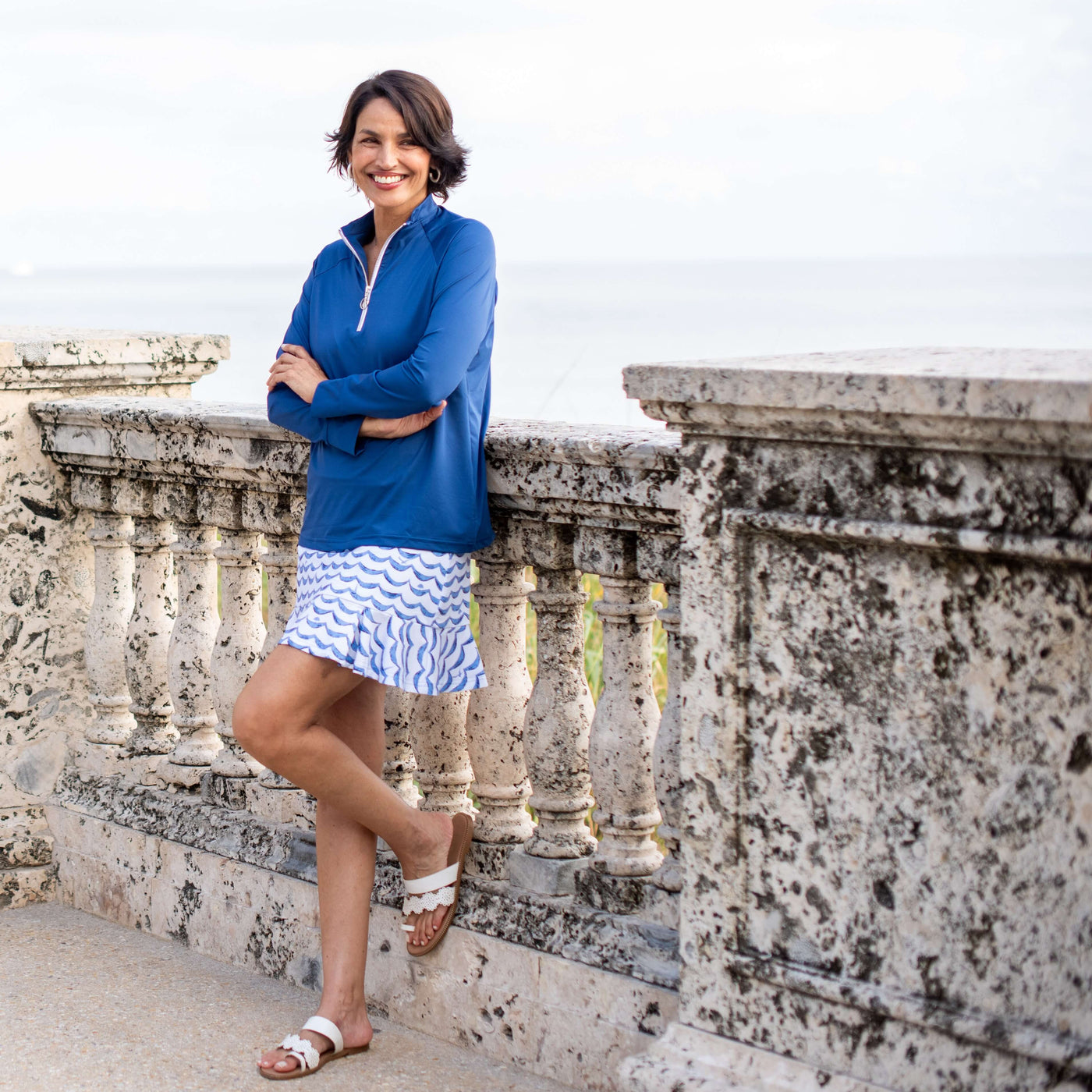 Woman in a blue top and patterned skirt standing on a stone balcony.