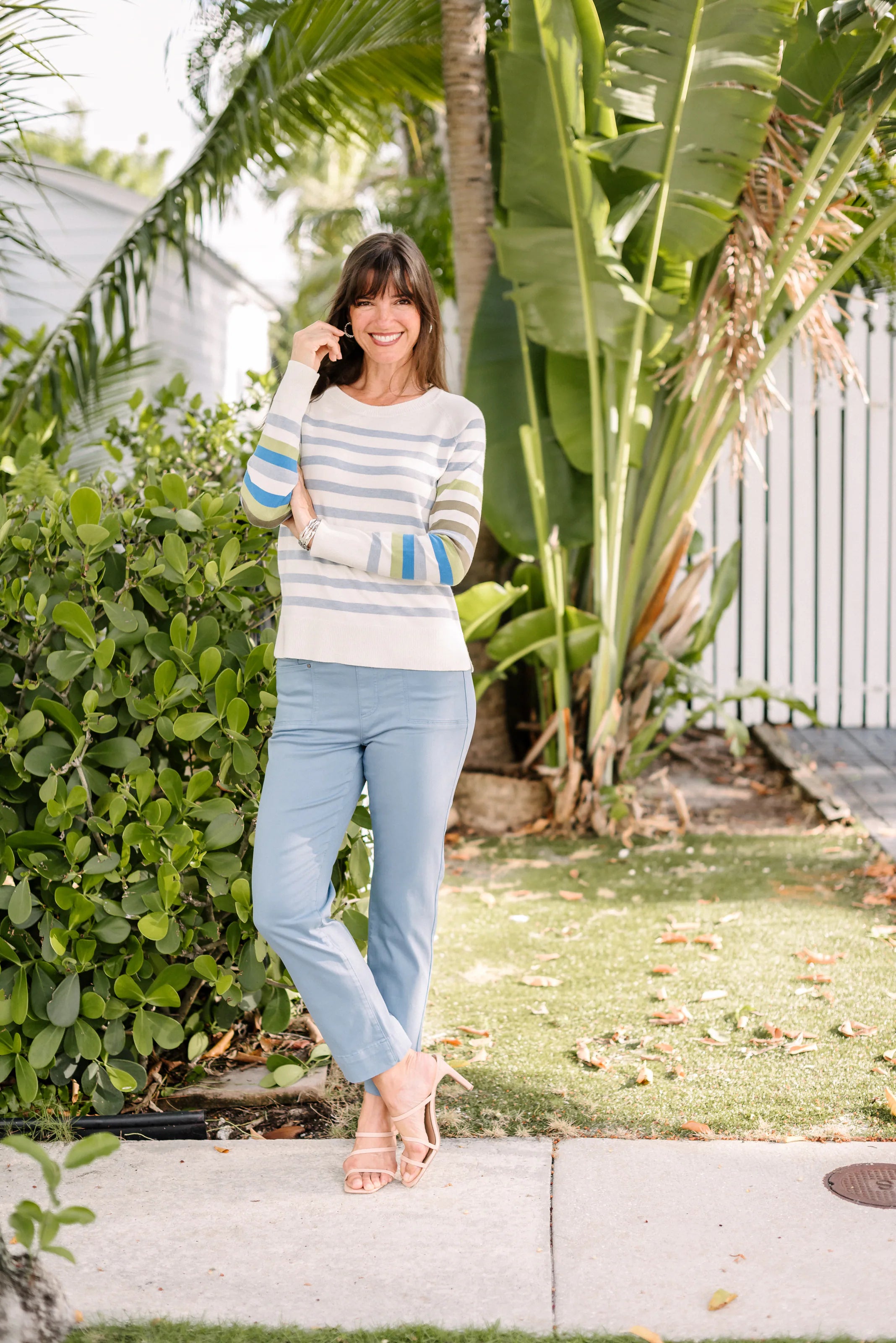 Woman in a pastel-striped sweater and pale blue pants beside tropical foliage.