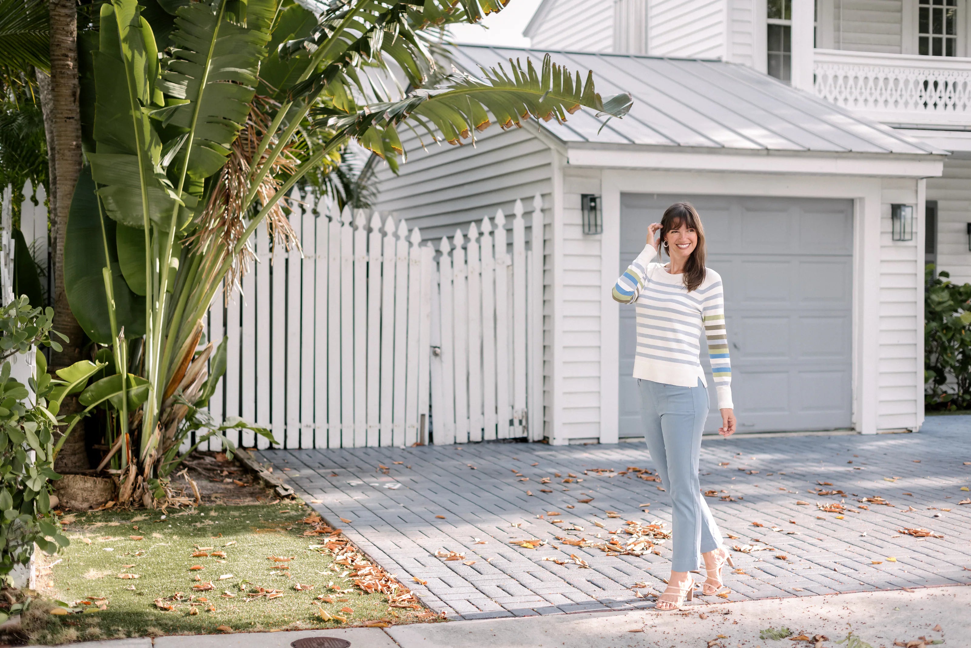 Full-length view of a woman in a striped sweater and pale blue pants outside a white coastal house.