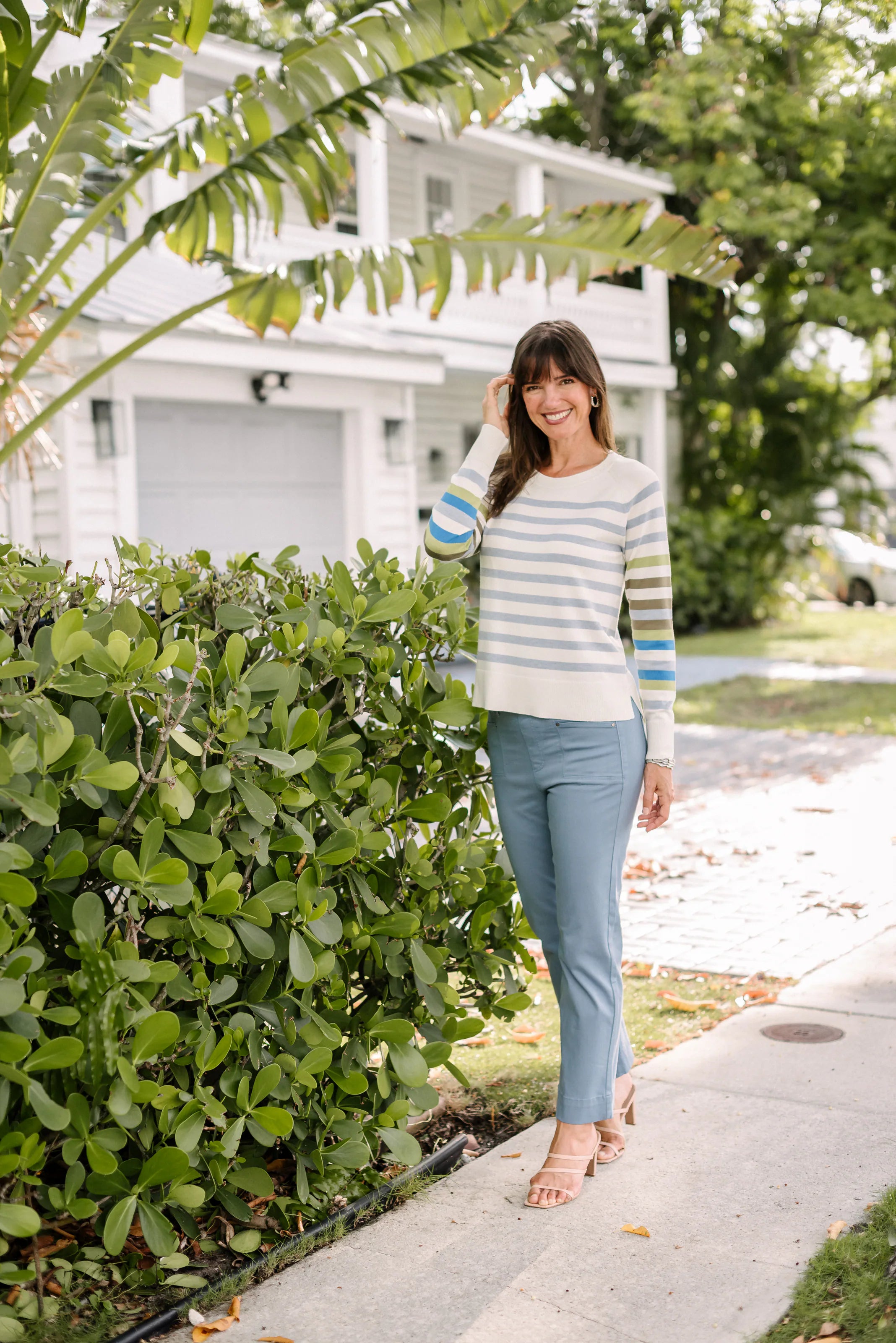Woman smiling in a pastel-striped sweater and pale blue pants outside a coastal house.