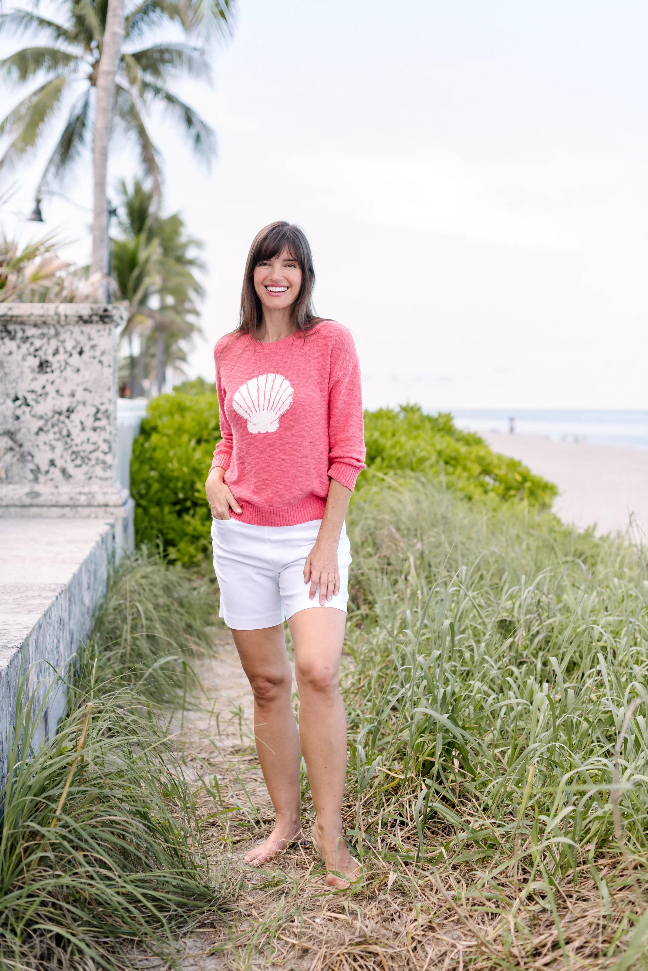 Woman standing in a pink short-sleeved sweater and white shorts near the beach.