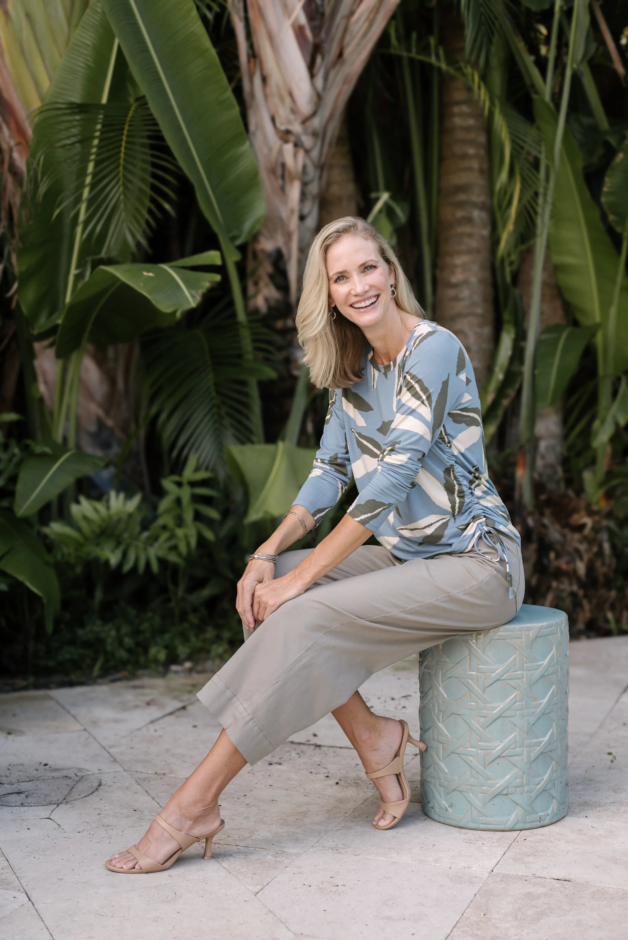 Woman seated in a blue-green leaf-print blouse and neutral pants.