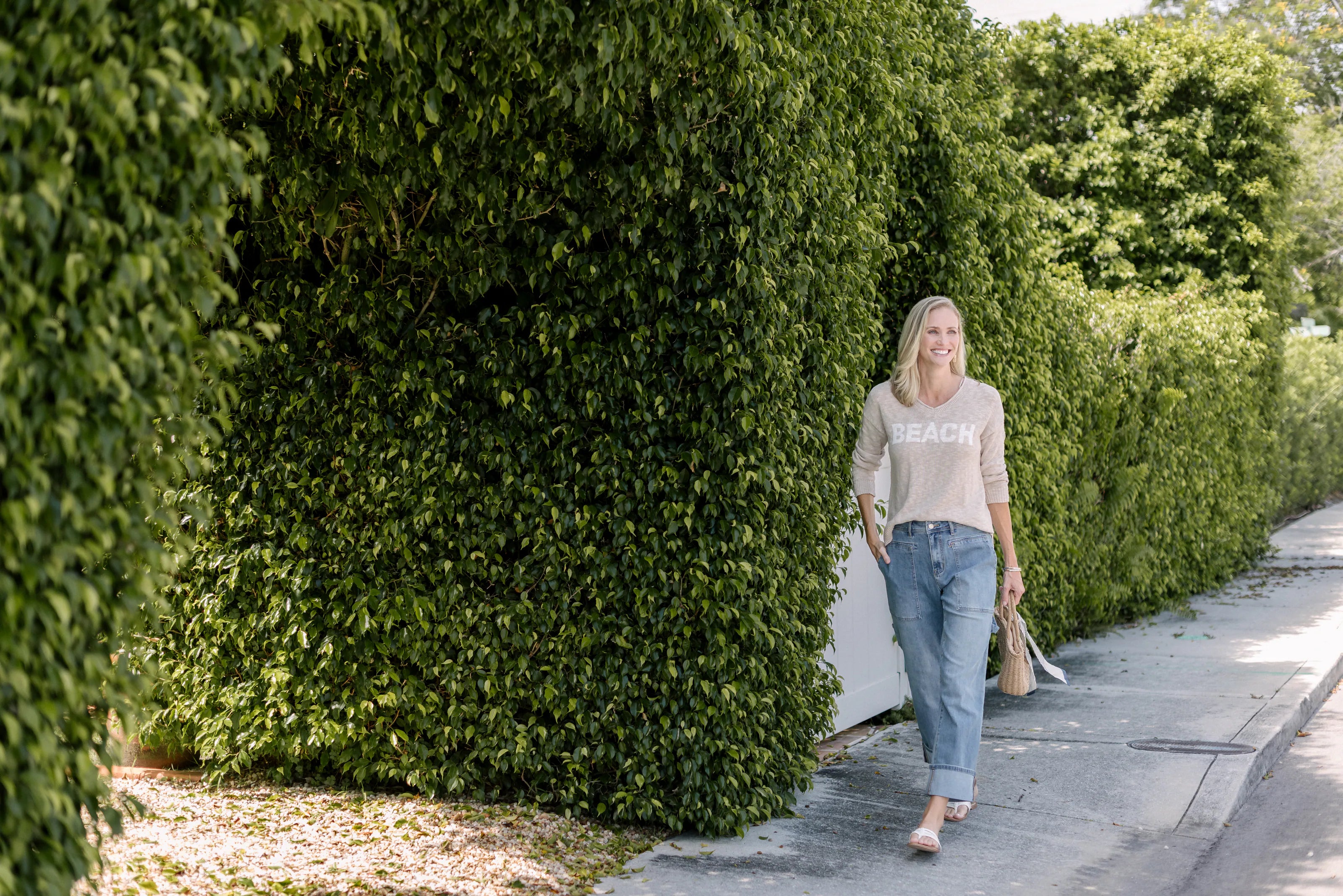 Woman wearing a taupe BEACH sweater and jeans beside a green hedge.